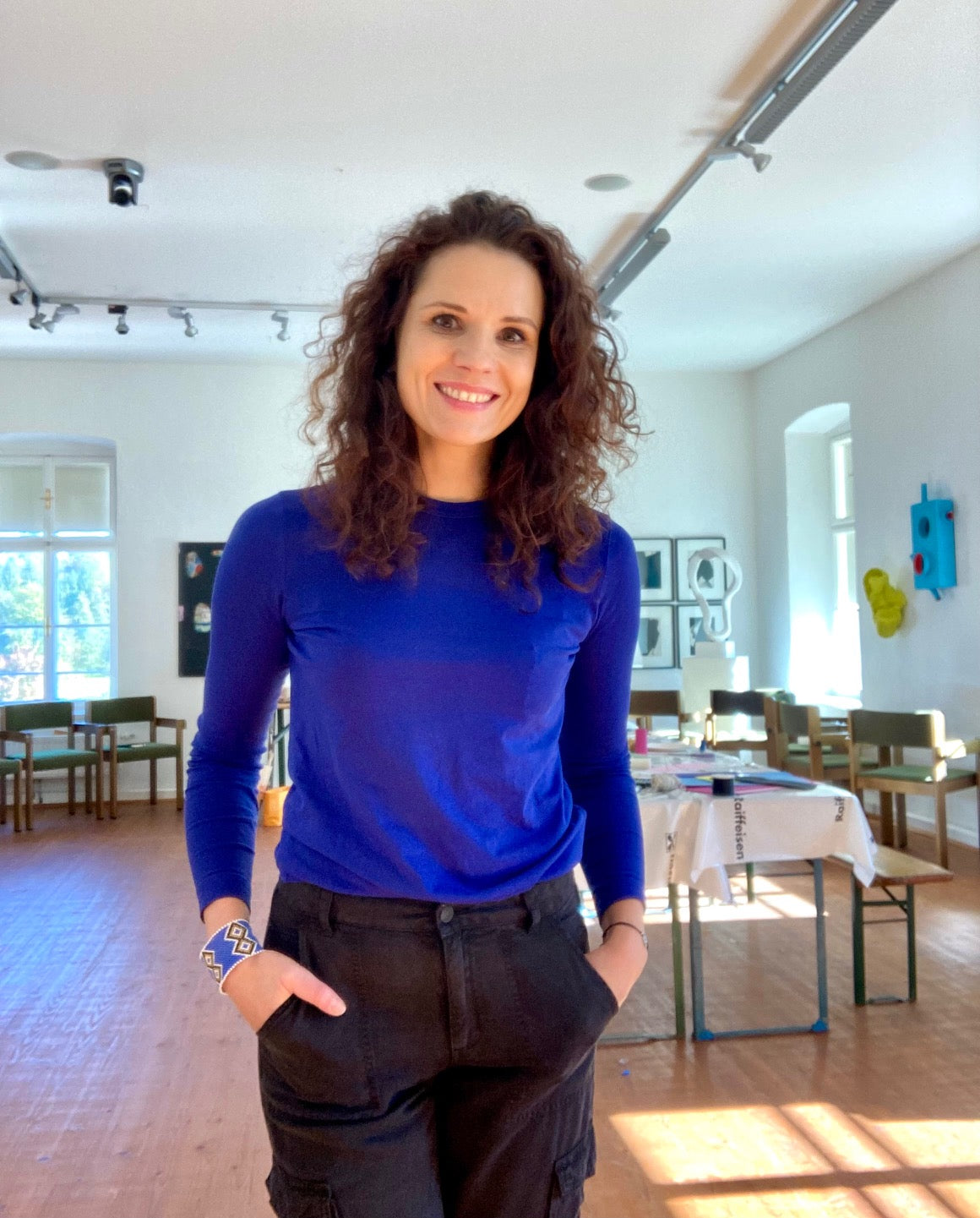 a woman with dark curly hair, wearing a blue T-shirt and black cargo pants standing in an atelier