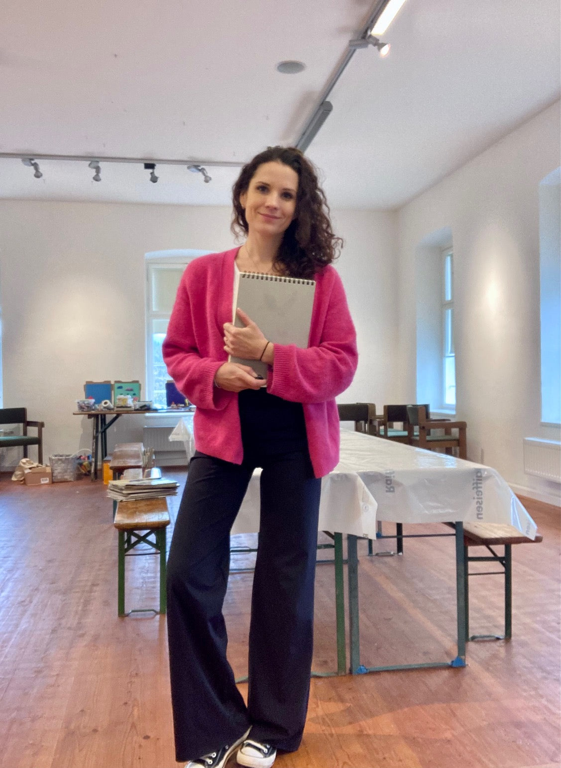 a woman with brown curly hair, wearing pink cardigan and dark blue trousers standing in an atelier